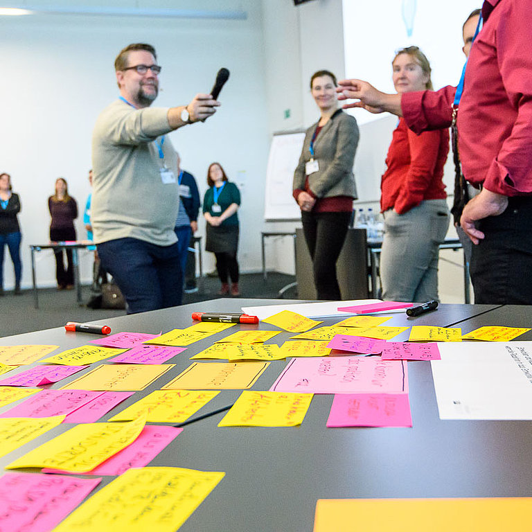 Workshop Tisch mit vielen Notizen, Menschen im Kreis, Mann reicht Mikrofon weiter